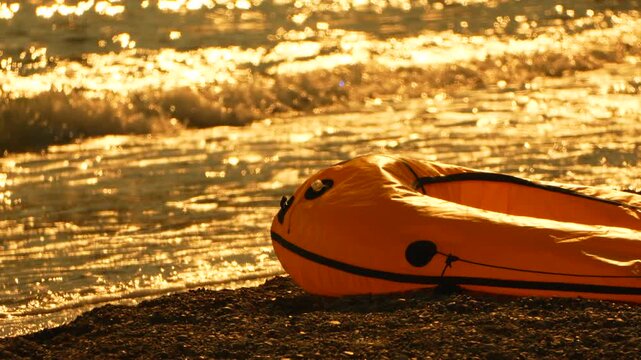 Inflatable yellow dinghy rests upon a dark gravel shoreline as bright tide waves roll in. Stationary long shot.