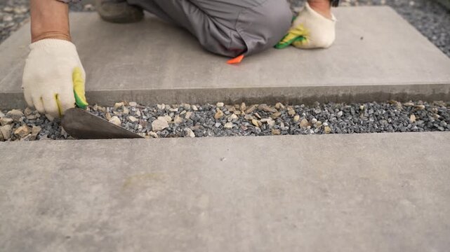 Closeup of gloved hand placing gray concrete pavers over crushed stone base. Natural daylight highlights texture contrast between smooth slabs rough gravel