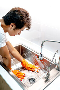 Asian male housekeeper with orange rubber glove scrubbing on kitchen sink surface with soap suds spreading around. Cleaning stainless steel sink for hygiene. Housework chores in the kitchen