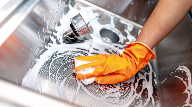 Housekeeper hand wearing orange rubber glove scrubbing on kitchen sink drainer with soap suds spreading around. Cleaning stainless steel sink for hygiene. Housework chores in the kitchen