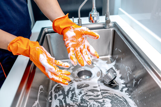 Housekeeper showing hands with orange rubber glove full with soap suds while scrubbing on kitchen sink surface. Cleaning stainless steel sink for hygiene. Housework chores in the kitchen