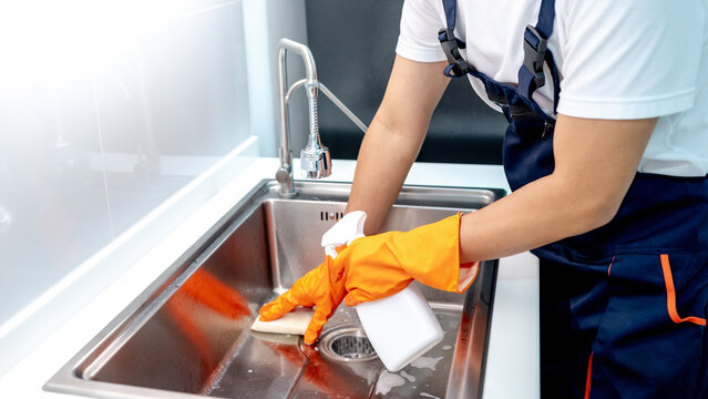 Male housekeeper wearing orange rubber gloves holding detergent spray bottle while scrubbing stainless steel kitchen sink with sponge. Home hygiene maintenance service