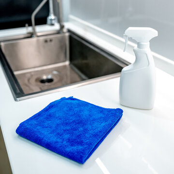 Blue microfiber cloth and detergent spray bottle on modern white countertop near stainless steel sink in domestic kitchen. Cleaning tool product for housework chores and home hygiene
