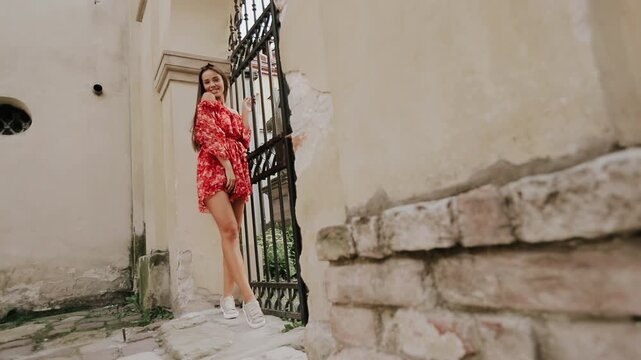 Young woman in red floral off-shoulder dress and white sneakers moving through ornate iron gate, one hand on bars, in worn urban courtyard with stone, brick, plaster and soft daylight, romantic