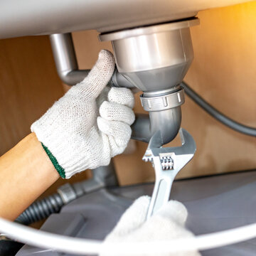 Plumber hand with glove fixing pipe connection with adjustable wrench and checking grease trap box under kitchen sink. Handyman repairing plumbing system in wooden cabinet.