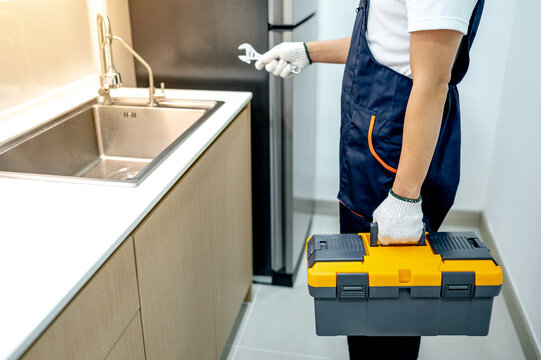 Male plumber with gloves holding yellow toolbox and wrench in the kitchen. Handyman in uniform prepares to fix plumbing system under stainless steel sink in wooden cabinet. Home maintenance service