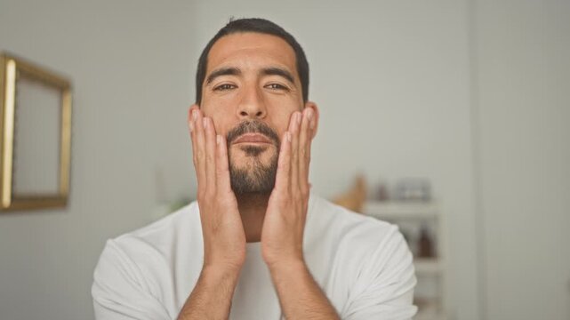 Young hispanic man touching and massaging his beard and jawline with both hands while checking skin in a studio mirror setting; confidence grooming.