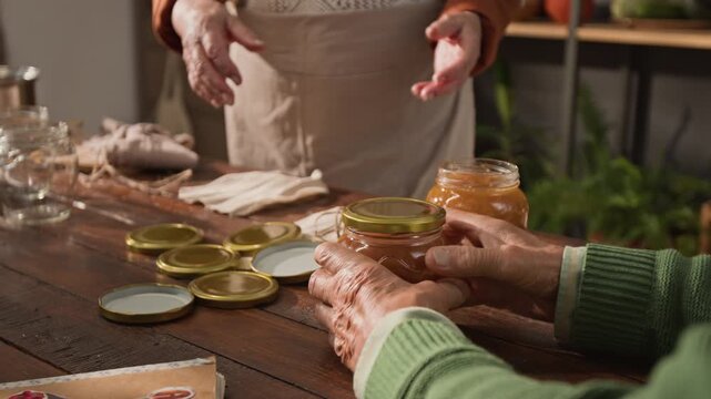 Unrecognizable mature man closing jar of homemade jam while senior woman wraps another jar in background. Ideal for promoting local organic food brands, artisanal products or retirement hobbies