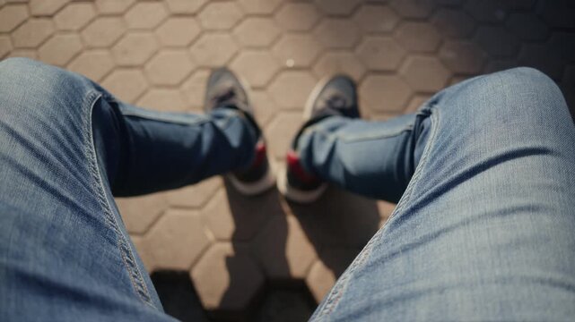 First person view of person sitting on pavement wearing blue jeans and sneakers.