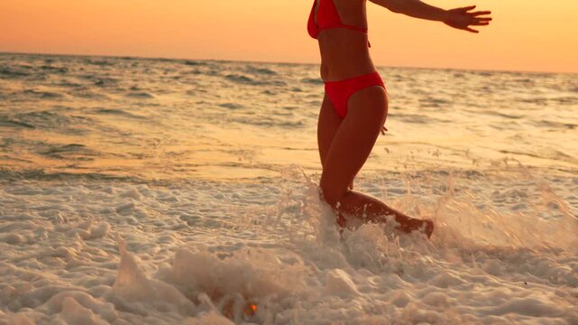 Red bikini bottom woman walks through shallow ocean waves at sunset along the coastline. Low angle tracking shot.