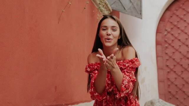Smiling young woman moves through narrow terracotta courtyard, giving affectionate kiss gesture while wearing red floral off-shoulder dress, ruffled bodice, cinched waist, warm sunlight