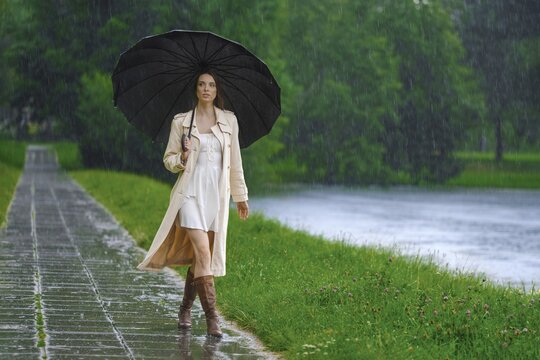 Woman walks along pathway beside river in park during rainy weather. She holds black umbrella and wears light-colored outfit