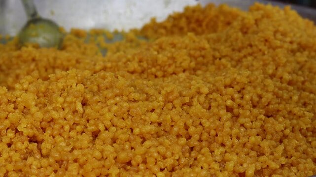 Close-up macro shot of freshly prepared sweet golden-orange boondi, a traditional Indian dessert and festival prasad, piled in a large metal container.
