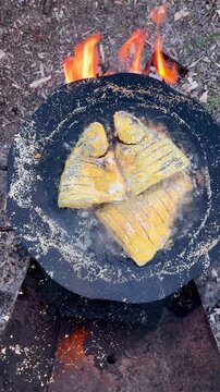 Golden cornmeal fried fish pieces sizzling over an open fire griddle