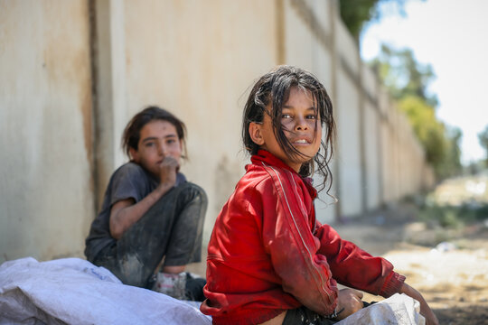 Thoughtful poor children sitting near a wall. Emotional portrait of a young refugee girl with sad eyes. Two Syrian girls in torn clothes sit by the roadside after rummaging through garbage for plastic