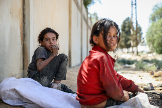 Thoughtful poor children sitting near a wall. Emotional portrait of a young refugee girl with sad eyes. Two Syrian girls in torn clothes sit by the roadside after rummaging through garbage for plastic