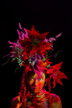 Conceptual fashion portrait of a young Southeast Asian woman wearing a crown made of artificial flowers with colorful tika powders on her face, shot with a hard light flash inside a studio