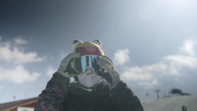 A person wearing a playful frog hat and rabbit face mask adjusts their ski goggles on a sunny day. They prepare for winter sports activities in the snowy mountains.