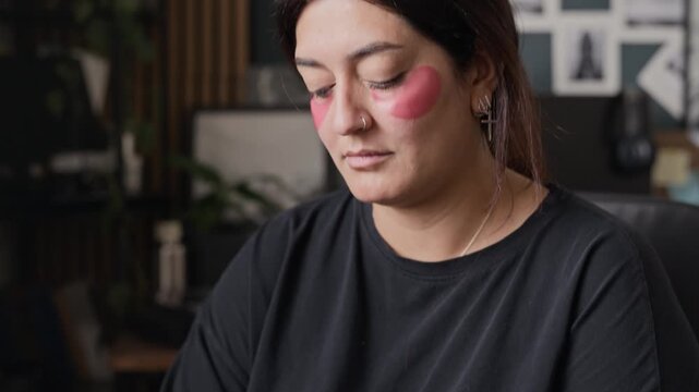 Young adult woman with pink eye patches holding cosmetic bottle, camera tilting up to her face. Ideal for skincare, self-care, and morning routine commercial campaigns