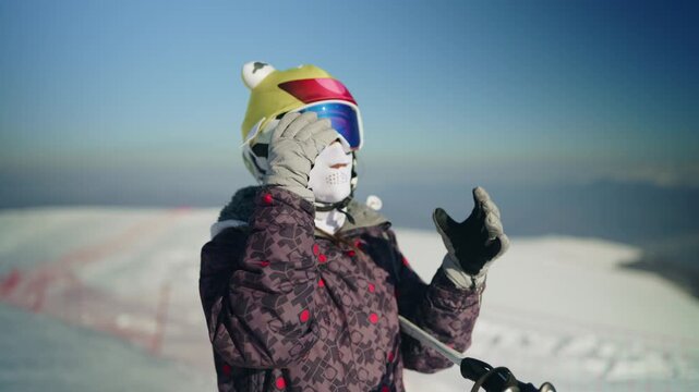 A young woman wearing a ski helmet and goggles adjusts her eyewear on a snowy mountain. She prepares for a day of skiing under a clear blue sky.