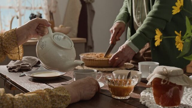 Hands of senior woman pouring tea from teapot into cup while senior man cutting pie in background. Perfect for retirement lifestyle and senior care commercial advertisements