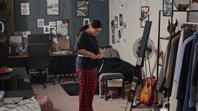 Young adult woman measuring waist with tape measure in front of mirror in bedroom. Video suitable for body positivity campaigns, weight loss programs, or fitness apps