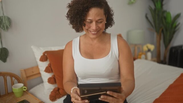 Woman in a white tank top holding a framed photo with both hands and smiling while seated on a bed in a sunlit bedroom; nostalgic memory.