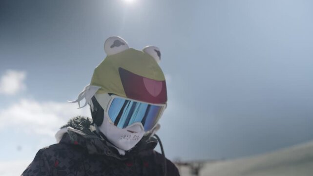 A person in a playful frog hat and ski goggles adjusts their winter gear. They are ready for snow sports on a bright, sunny mountain.