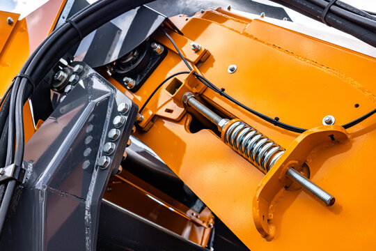 Heavy agricultural machinery tension spring and hydraulic hoses mechanism. Industrial equipment detail with threaded rod, steel bolts, pillow block bearing, and yellow metal structure.