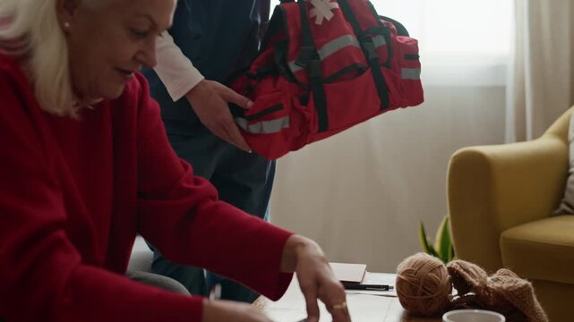 Young adult Caucasian male nurse carrying red medical bag, sitting on sofa next to senior Caucasian female patient to discuss health. Video suitable for geriatric care commercial