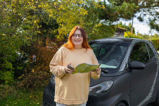 Thoughtful young caucasian woman holding a paper guide map and checking a smartphone navigation app while standing next to her car during a road trip.