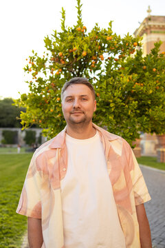 Medium shot of a positive caucasian man in a pink shirt and white t-shirt looking at the camera and smiling, standing against a background of green trees and sunny backyard