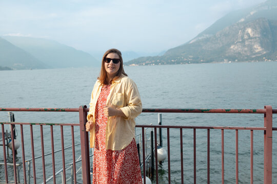 Beautiful millennial woman in casual dress, sunglasses and sneakers posing on a wooden pier or promenade at a scenic European travel destination.