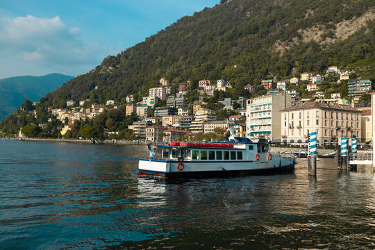 A breathtaking wide shot of Lake Como (Lago di Como) in Lombardy, Italy. The scene showcases vibrant blue water with moving boats, charming coastal town architecture nestled on hills, and green