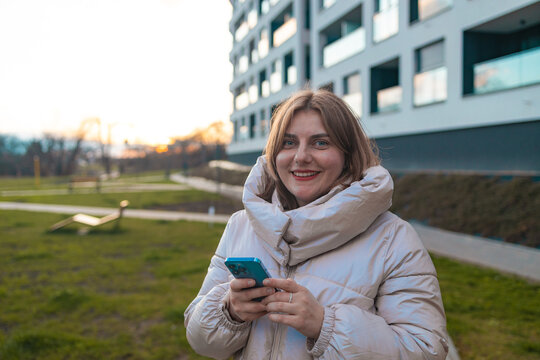 A woman is smiling and looking at the camera in a grassy outdoor area with a modern building in the background. The scene conveys a happy and relaxed mood, with natural green and white hues.