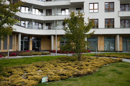 A beautiful scenic view of a modern residential apartment building or alpine style resort hotel. The property features well-manicured green lawns, decorative bushes, and landscaped gardens in the