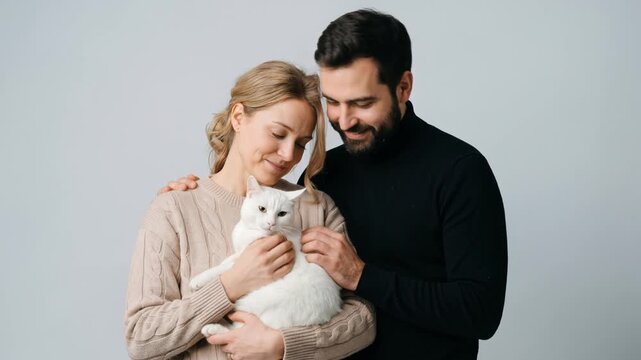 Couple holding white cat on grey background. Man and woman stroking domestic pet in studio. Pet owner love and animal care concept