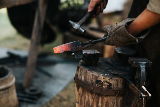 A blacksmith works with hot metal using tongs and hammers in a medieval festival. The activity takes place in a historical setting with tools and equipment typical of the time