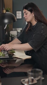 Vertical video of young adult woman slicing cucumber on cutting board and writing in notebook. Clip is suitable for healthy lifestyle, meal planning, and diet tracking commercials