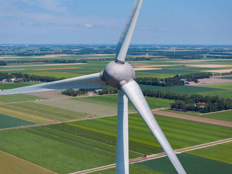 Aerial view of a large white wind turbine standing amidst a vast expanse of green agricultural fields and patchwork farmland in the Netherlands.
