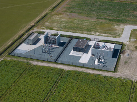 Aerial view of an electrical substation with transformers and switchgear equipment enclosed by a fence, situated in the middle of agricultural fields in the Netherlands.