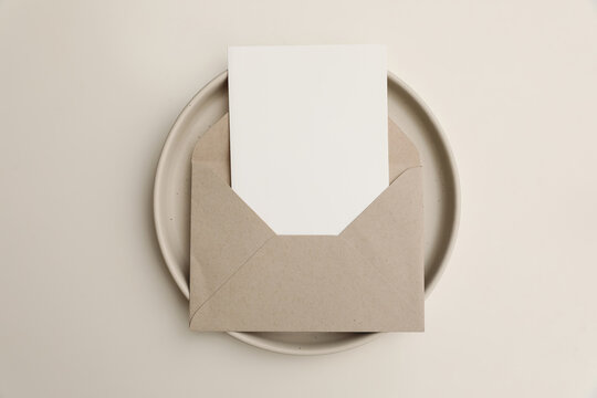 Blank greeting card mockup in open kraft envelope on ceramic plate. Minimalist stationery scene with copy space, neutral beige table background for invitation, branding, wedding design. Flatlay top