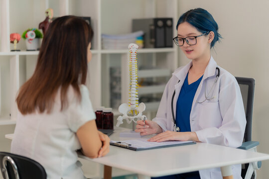 Young Asian female doctor or nurse with blue hair explaining dosage instructions for medication bottle to a senior female patient in a hospital or clinic.