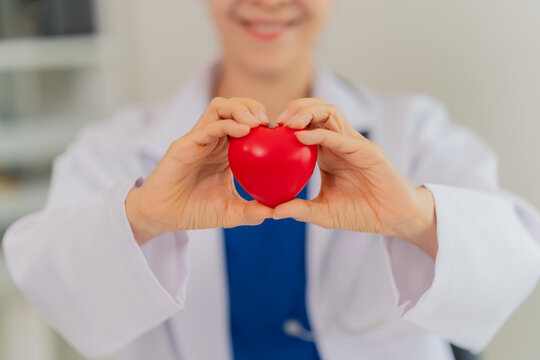 Smiling senior female doctor holding red heart model during online medical consultation on laptop.
