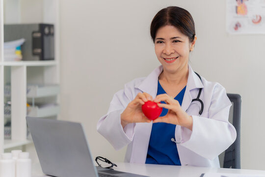Smiling senior female doctor holding red heart model during online medical consultation on laptop.