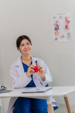 Smiling senior female doctor holding red heart model during online medical consultation on laptop.