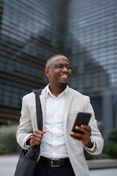 CFO, black businessman, using smartphone, calling, outdoor near office building, vertical. African american business man smiling, committed to career growth, building strong client partnerships.