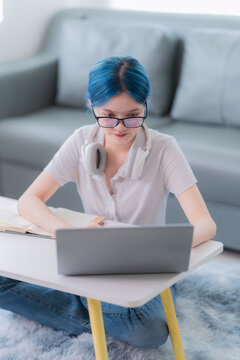 Young Asian woman with blue hair wearing headphones studying online with laptop and writing notes at home.