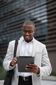 Financial analyst black businessman outdoor near office building, using tablet, swiping screen, vertical. African american business man smiling, staying connected with technology, managing accounts.