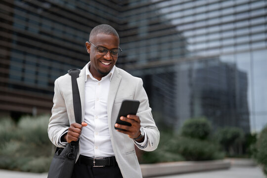 Vice president african businessman outdoor near office building, using smartphone, scrolling news. Black business man smiling, managing work remotely, building strong client partnerships. Copy space